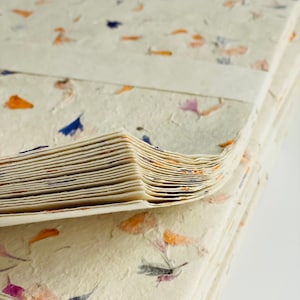 May include: A stack of handmade paper with embedded dried flowers. The paper is a light cream color with orange, purple, and blue flowers.