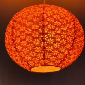 Può includere: Una lanterna rotonda di carta arancione con un motivo floreale. È sospesa a un filo sottile ed emette una calda luce invitante. Il design presenta piccole forme floreali ripetute, creando un effetto delicato e decorativo. La lanterna è un elemento di illuminazione decorativo.