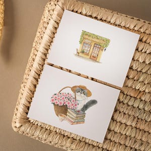 May include: Two watercolor art prints on white paper. One depicts a small shop with a green awning. The other shows a cat in a hat with a basket of pink roses, a stack of books, and a teacup. The prints are on a woven basket.