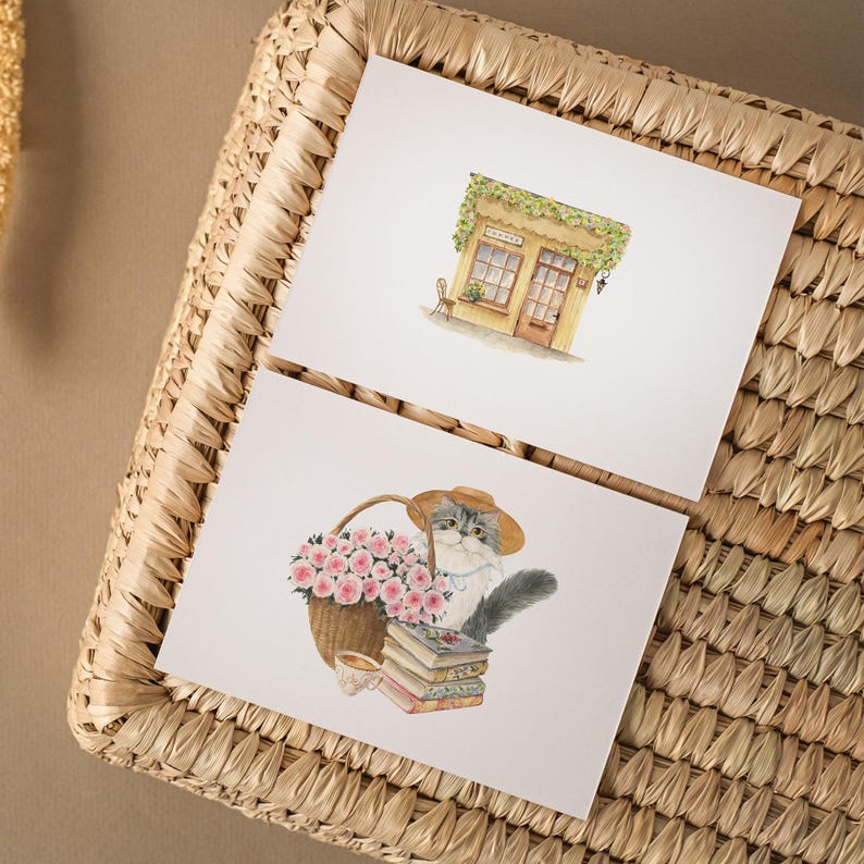 May include: Two watercolor art prints on white paper. One depicts a small shop with a green awning. The other shows a cat in a hat with a basket of pink roses, a stack of books, and a teacup. The prints are on a woven basket.