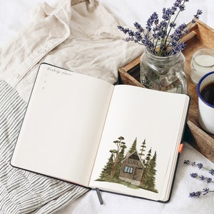 May include: Open notebook with a watercolor illustration of a small cabin in the woods on the right page. The left page has the words "Weekly Plans" written at the top and a list of numbers underneath. The notebook is on a white bedspread with a white shirt, a wooden tray with a cup of coffee, a candle, and a vase of lavender.