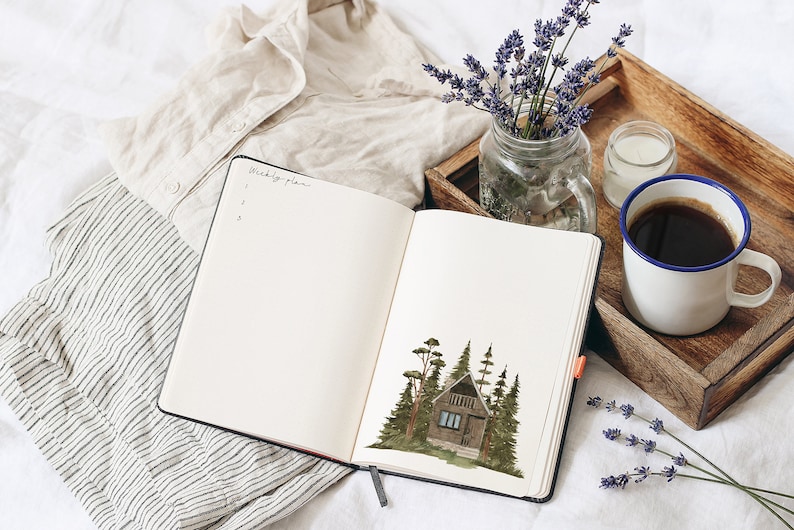 May include: Open notebook with a watercolor illustration of a small cabin in the woods on the right page. The left page has the words "Weekly Plans" written at the top and a list of numbers underneath. The notebook is on a white bedspread with a white shirt, a wooden tray with a cup of coffee, a candle, and a vase of lavender.