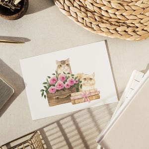 May include: A watercolor illustration of two cats, one gray and one cream-colored, with pink roses. The cats are positioned near a wooden box and books tied with a pink ribbon. The image is on a white card.