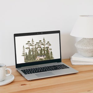 May include: A silver laptop computer with a white background on the screen. The background shows a watercolor illustration of a wolf walking through a forest of green trees. The laptop is sitting on a light brown wooden desk with a white ceramic mug and saucer to the left and a white lamp with a gray base on a stack of books to the right.
