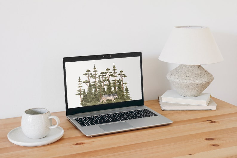 May include: A silver laptop computer with a white background on the screen. The background shows a watercolor illustration of a wolf walking through a forest of green trees. The laptop is sitting on a light brown wooden desk with a white ceramic mug and saucer to the left and a white lamp with a gray base on a stack of books to the right.