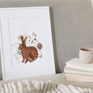May include: A framed print of a brown rabbit surrounded by watercolor flowers in shades of blue, orange, and yellow. The print is on a white background and is framed in a white frame.