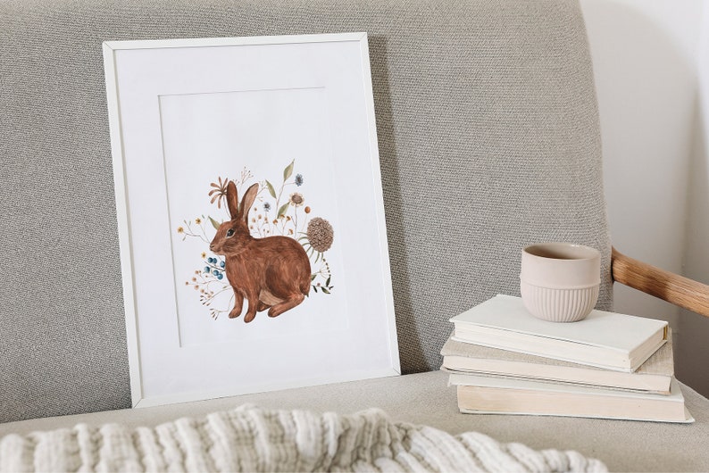 May include: A framed print of a brown rabbit surrounded by watercolor flowers in shades of blue, orange, and yellow. The print is on a white background and is framed in a white frame.