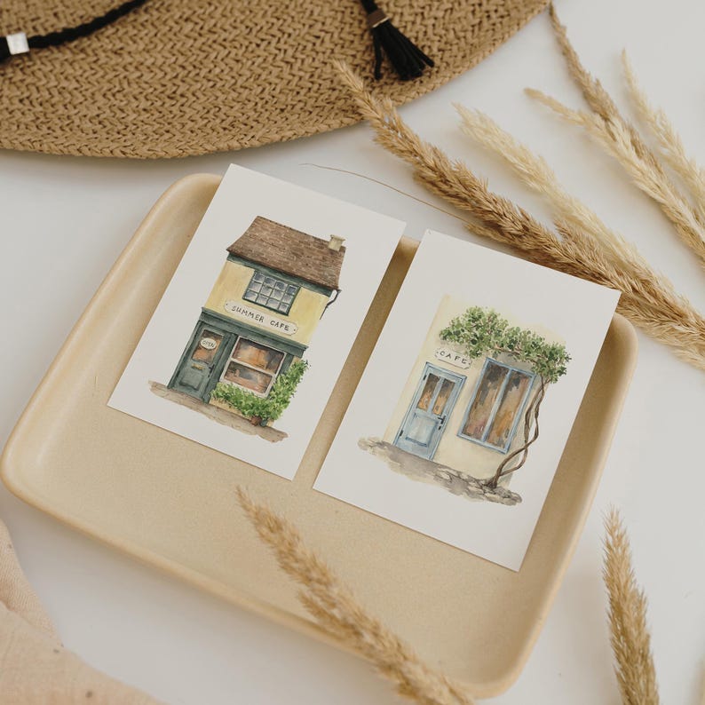May include: Two watercolor postcards featuring quaint cafe storefronts. One card shows a yellow building with the text "SUMMER CAFE" above the door. The other card shows a cafe with the text "CAFE" above the door. The cards are on a beige tray with wheat stalks.