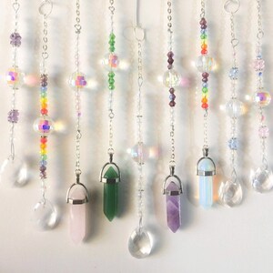 May include: A set of nine crystal and gemstone pendants hanging from silver chains. The pendants include clear crystal, amethyst, rose quartz, green aventurine, and opal. Each pendant is adorned with beads in various colors, including rainbow, green, and purple.