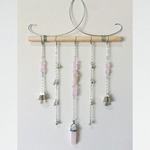 May include: A pink crystal and silver bell wind chime with a wooden dowel and a metal frame. The wind chime features pink crystal beads and silver bells.