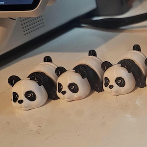 Könnte beinhalten: Fünf kleine schwarz-weiße Pandabären-Figuren. Die Pandas liegen auf dem Bauch mit dem Kopf zur Seite gedreht. Sie sind aus weißem Material mit schwarzen Details bemalt.