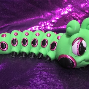 Puede incluir: Una figura de oruga verde con detalles en rosa y negro. El juguete tiene ojos grandes y expresivos y un cuerpo segmentado. El fondo es una tela morada brillante.