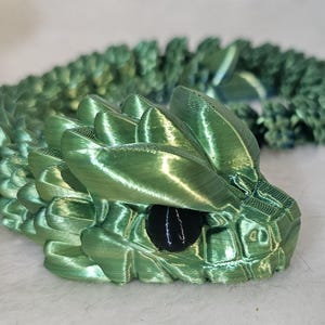 May include: A green 3D printed dragon figurine with a textured, scaly surface and black eyes. The dragon is curled up in a coil shape.