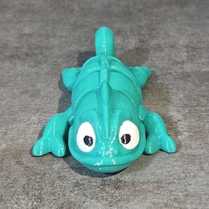 May include: A teal green chameleon figurine with white eyes and a textured surface. The chameleon is posed with its legs outstretched and its head tilted slightly upwards.