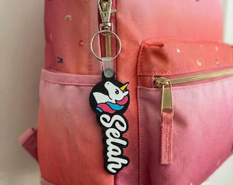 May include: A pink and red ombre backpack with a gold zip and a unicorn keyring. The keyring is black with a unicorn graphic and the name "Selah" in white script. The backpack has a top handle and a front pocket.