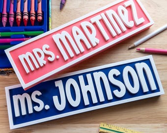 May include: Two rectangular nameplates, one pink and one blue, with white lettering. The pink nameplate reads "MRS. MARTINEZ", and the blue one reads "MS. JOHNSON". Crayons, coloured pencils, and a ruler are scattered around the nameplates.
