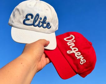 May include: A white baseball cap with the word Elite embroidered in blue script and a red baseball cap with the word Dingers embroidered in white script. Both caps feature a structured crown, a curved brim, and a button on the top center.