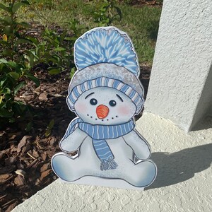 Puede incluir: Un recorte de muñeco de nieve blanco con un sombrero a rayas azul y blanco, un pompón azul y una bufanda a rayas azul y blanco. El muñeco de nieve tiene una nariz roja y ojos negros.