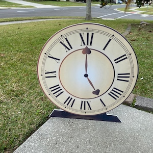 May include: A large, round clock with Roman numerals and black hands pointing to 5 o'clock. The clock face is cream-colored with a brown border and black markings. It is standing upright on a concrete surface.