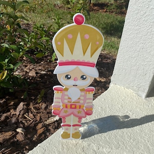 Puede incluir: Una figura decorativa de cascanueces independiente. La figura tiene un uniforme rosa y dorado, una barba blanca y una corona. La figura está de pie sobre una superficie blanca, con un fondo de vegetación y un cielo soleado.