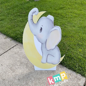 Puede incluir: Un elefante gris con una luna creciente amarilla detrás. El elefante está sentado en la luna y tiene la trompa levantada. La imagen es un recorte para una decoración de fiesta.