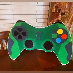 May include: A green and black cardboard cutout of a video game controller. The controller has four buttons, a directional pad, and two triggers.