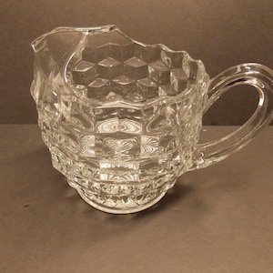 May include: A clear glass pitcher with a diamond pattern and a handle. The pitcher is made of thick glass and has a slightly chipped rim.