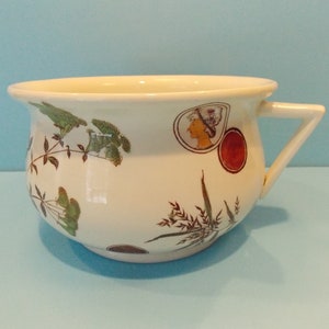 May include: White ceramic chamber pot with a floral and botanical design. The pot has a single handle and a rounded shape.