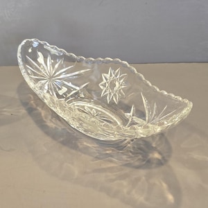 May include: A clear, oval-shaped glass dish with starburst and etched designs. The dish has a scalloped edge and is likely used for serving or display. The glass is transparent, allowing light to pass through.