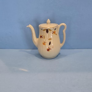 May include: Cream-colored vintage teapot with a lid and handle. The teapot features a floral design in shades of orange, brown, and yellow. Gold trim accents the handle and spout. A classic piece for serving tea.