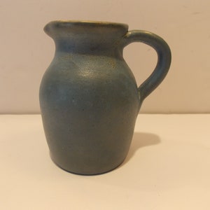 May include: A small, blue ceramic pitcher with a rounded body and a handle. The pitcher has a matte finish.