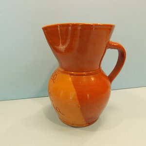 May include: A terracotta pitcher with a wide mouth and a handle. The pitcher is decorated with a brown glaze and has a light brown stripe down the side.