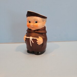 May include: A brown ceramic figurine of a monk with a white rope belt and a white hat. The monk has a round face with a smile and large eyes.