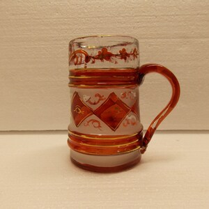 May include: A glass beer stein with a white frosted finish and red and gold painted designs. The stein has a handle and a rounded base.