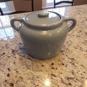 May include: A light blue ceramic bean pot with a lid and two handles. The pot is sitting on a granite countertop.