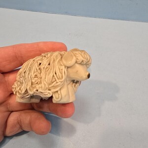 May include: A small, white ceramic sheep figurine with a curly, detailed wool design. The sheep is standing on its four legs and has a black eye.
