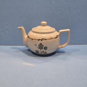 May include: A cream-colored ceramic teapot featuring a lid with a small knob, a curved spout, and a handle. The body is decorated with a horizontal brown line pattern featuring leaves and a small illustration of a birdhouse and a topiary tree.