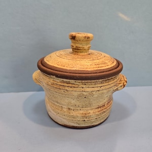 May include: A small, cream-colored ceramic pot with a lid and two handles. The pot has a brown rim and a speckled glaze.