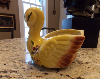 May include: A yellow ceramic swan planter with brown and green painted details. The planter has a wide opening for planting flowers or other greenery.