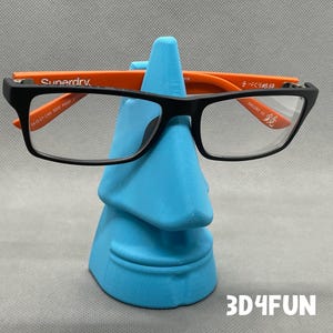 May include: A blue plastic nose-shaped stand with a pair of black framed glasses with orange arms resting on it. The glasses have the text "Superdry." on the arms. The stand has the text "3D4FUN" printed on the surface.