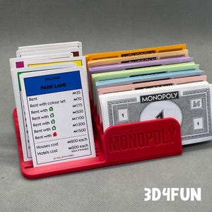 May include: A red Monopoly game property card holder with a Monopoly game board card for Park Lane. The card holder has the word "Monopoly" printed on the front. The card holder is holding a stack of Monopoly money cards.