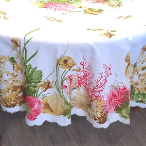 Op de afbeelding: Een ronde witte tafelkleed met geschulpte rand, met een kleurrijk onderwaterontwerp. Het patroon bevat koraal, zeegroenten en kwallen in tinten roze, groen en geel. Het tafelkleed is gedrapeerd over een houten tafel.