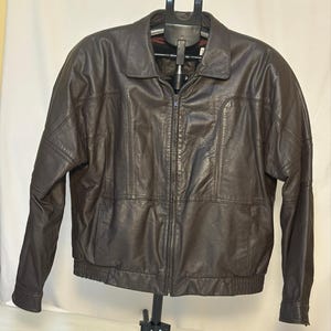 May include: A dark brown leather jacket featuring a full-length front zipper, a standard collar, and long sleeves with cuffs. The jacket includes vertical seam detailing across the chest and a ribbed elastic waistband for a structured fit.
