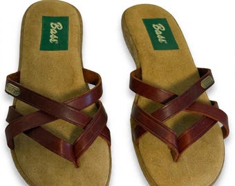 Könnte beinhalten: Ein Paar braune Ledersandalen mit überkreuzten Riemen und Zehensteg. Das Fußbett ist hellbraun und mit einem grünen rechteckigen Etikett versehen, das den Schriftzug Bass in Weiß zeigt. Ein kleines Bass-Logo aus goldfarbenem Metall sitzt am Riemen.