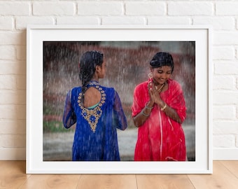 Puede incluir: Una fotografía enmarcada que muestra a dos personas bajo la lluvia. Una viste una prenda azul de manga larga con bordados dorados y espalda descubierta, mientras la otra lleva una prenda roja con joyas doradas y manos unidas en oración.
