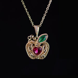 May include: A gold-coloured apple pendant necklace featuring a heart-shaped red gemstone. The apple design includes a green leaf and is suspended from a delicate chain. The background is black.
