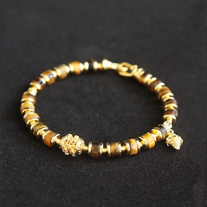 May include: A gold-colored bracelet with tiger eye beads and gold accents. The bracelet features a decorative gold bead with red accents and a small charm. The bracelet is on a black background.