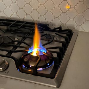 以下が含まれることがあります： 黒い鋳鉄製の五徳を備えたステンレス製のガスコンロ。一つのバーナーが明るい青とオレンジ色の炎で燃焼しています。コンロの奥には、光沢のある白いアラベスク模様のタイル壁が設置されており、光が反射しています。