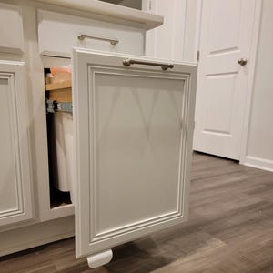 May include: White kitchen cabinet with a pull-out bin. The cabinet door is open and has a silver handle.