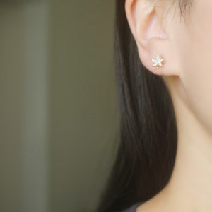 May include: A close-up of a gold-colored stud earring in the shape of a star, encrusted with small, clear gemstones. The earring is worn on an ear, with dark hair and a black top visible.